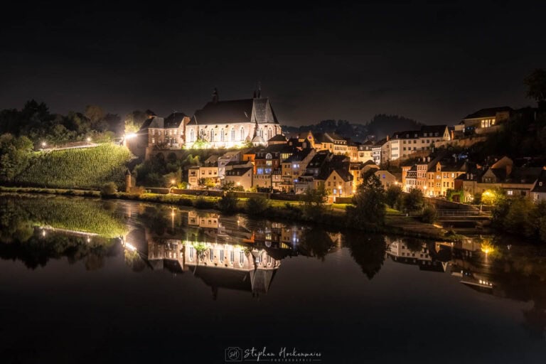 St. Laurentius und Altstadt von Saarburg bei Nacht mit Spiegelung in der ruhigen Saar