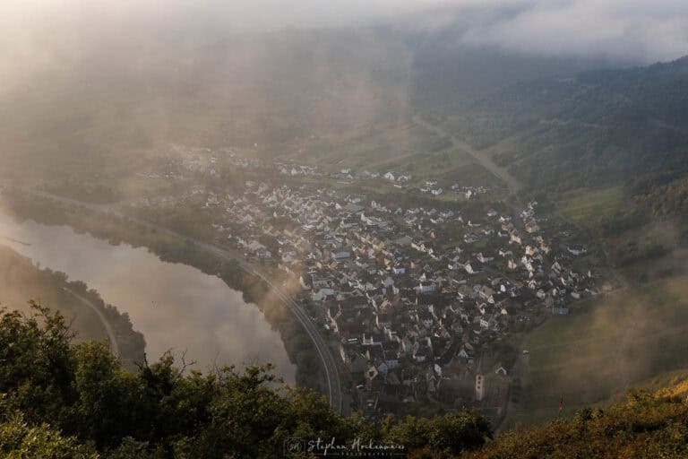 Blick vom Calmont Gipfelkreuz auf den Ort Bremm mit Nebel und Morgenlicht über dem Moseltal