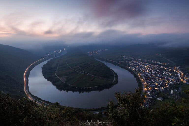 Moselschleife bei Bremm in der Blauen Stunde mit Nebel, beleuchtetem Ort und Lichtspuren entlang der Mosel